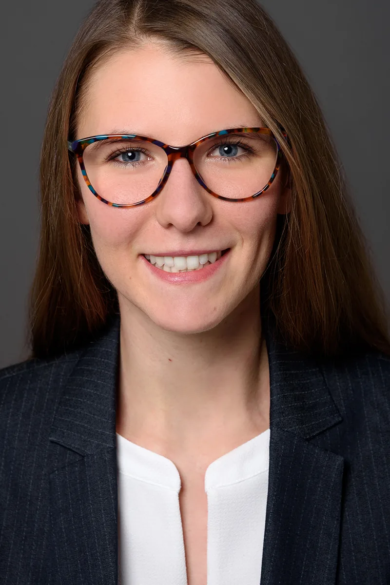 Laura Stess lächelnd, im dunklen Blazer vor grauem Hintergrund